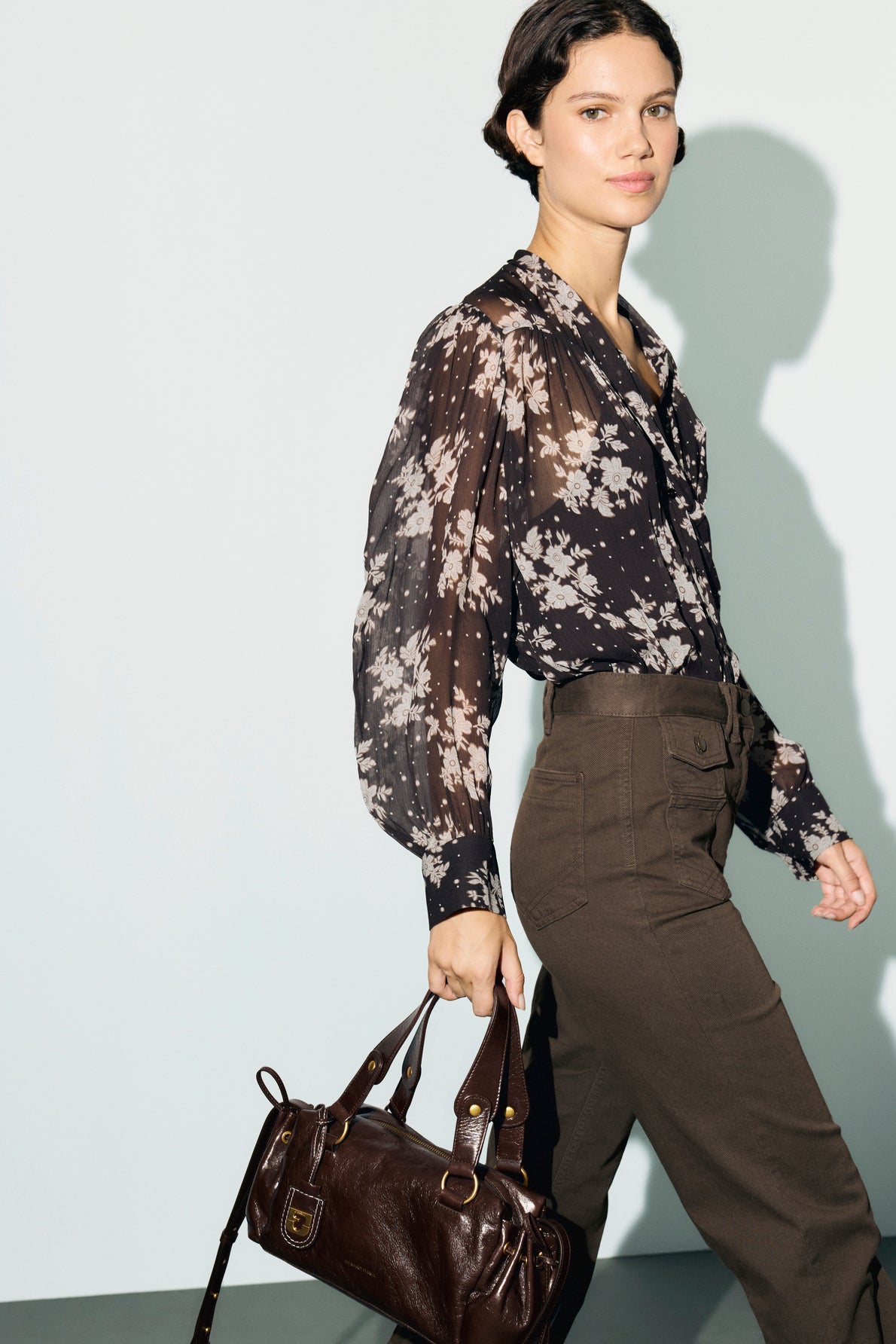 Woman with dark hair wearing a black chemise en mousseline à imprimé floral and olive green pants, holding a dark brown leather handbag, walking against a plain light background.