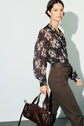 Woman with dark hair wearing a black chemise en mousseline à imprimé floral and olive green pants, holding a dark brown leather handbag, walking against a plain light background.