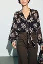 A person wearing a sheer black chemise en mousseline with an imprimé floral and a col lavallière, paired with high-waisted dark brown pants, stands against a light background, holding a dark bag.