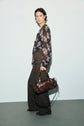 Woman with short dark hair stands against a light background, wearing a chemise en mousseline à imprimé floral noir transparent, wide-legged dark gray pants, and brown shoes. She holds a dark brown handbag in her left hand and looks towards the camera.