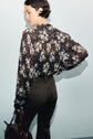 A person with short dark hair facing away, wearing a chemise en mousseline noire sheer with a white imprimé floral pattern and dark high-waisted pants, holding a dark bag against a light background.