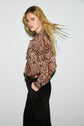 Person with long blonde hair wears a brown chemise cachemire featuring a paisley pattern, paired with dark pants and a belt. They stand sideways against a plain light background, looking at the camera with hair blowing back.