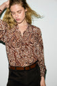 A person with long blonde hair wears a brown chemise cachemire, featuring a paisley pattern and tucked into black pants with a brown belt, standing against a light gray background.