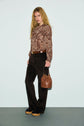 A person with long blonde hair wears a chemise cachemire—a paisley-patterned shirt crafted from flowing viscose haboutaï—paired with dark brown pants, platform shoes, and a brown handbag, standing against a plain pale background.