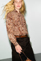 A person with long blonde hair wears a brown chemise cachemire paisley blouse, dark pants, and a brown belt, holding a brown handbag against a light background.