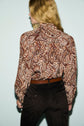 A person with long, blonde hair stands facing away, wearing a brown paisley-patterned chemise cachemire tucked into high-waisted dark pants with a brown belt.