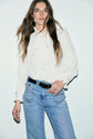 A person with long brown hair wears a white chemise à volants with a bow tie at the neck and light blue jeans, standing against a plain white background.