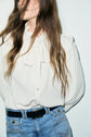 A person with long brown hair wears a cream chemise à volants with a bow tie and ruffled collar, tucked into high-waisted blue jeans with a black belt. The face is partially out of frame.