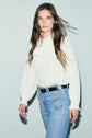 A woman with long brown hair wears a white chemise à volants and blue jeans with a black belt, standing against a light gray background.