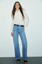 Woman stands against a plain background, wearing a chic chemise col montant with a bow at the neck, light blue high-waisted flared jeans, a black belt, and platform shoes. She has long, wavy brown hair.