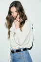 A woman with long brown hair wearing a cream chemise à volants and high-waisted blue jeans stands against a light background, looking at the camera with a slight smile and holding a strand of hair near her face.