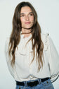 A person with long brown hair, wearing a cream chemise à volants and blue jeans, stands in front of a plain light background, looking slightly to the side and smiling gently.