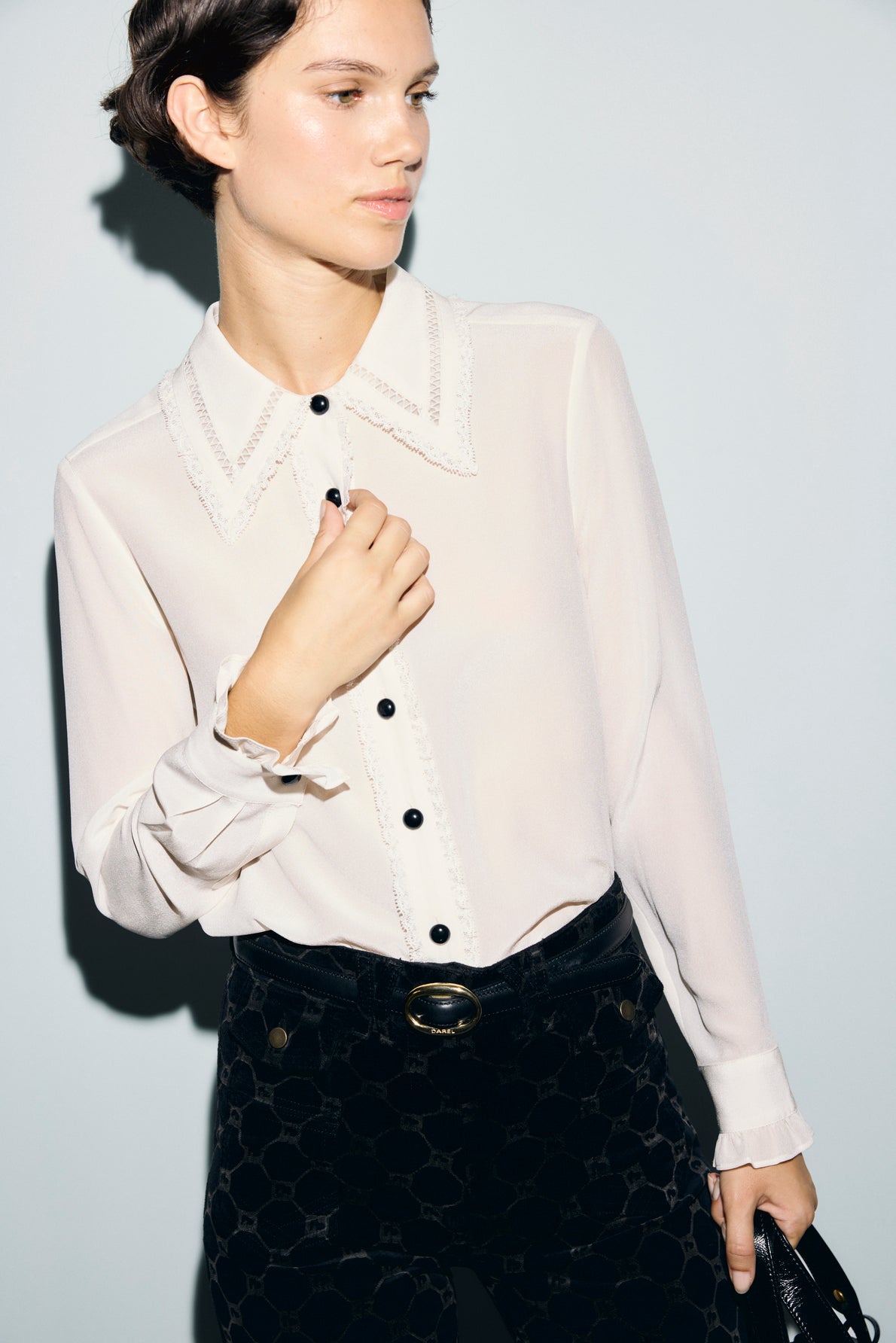Woman wearing a chic chemisier femme—a sheer white blouse with black buttons—paired elegantly with high-waisted black pants and a belt, posing gracefully against a light background.