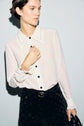 Woman wearing a chic chemisier femme—a sheer white blouse with black buttons—paired elegantly with high-waisted black pants and a belt, posing gracefully against a light background.