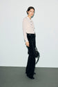 A person stands against a plain background wearing a chemisier femme blanc, buttoned up and paired with high-waisted black pants featuring a textured pattern, while holding a black handbag.