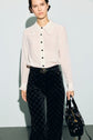 A woman wearing a white chemisier femme with a decorative collar and black patterned pants holds a black handbag. She stands against a light background, looking at the camera.