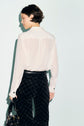 A person with short dark hair wearing a chemisier dentelle blanc transparent and black patterned pants stands facing away, holding a black handbag, against a light background.