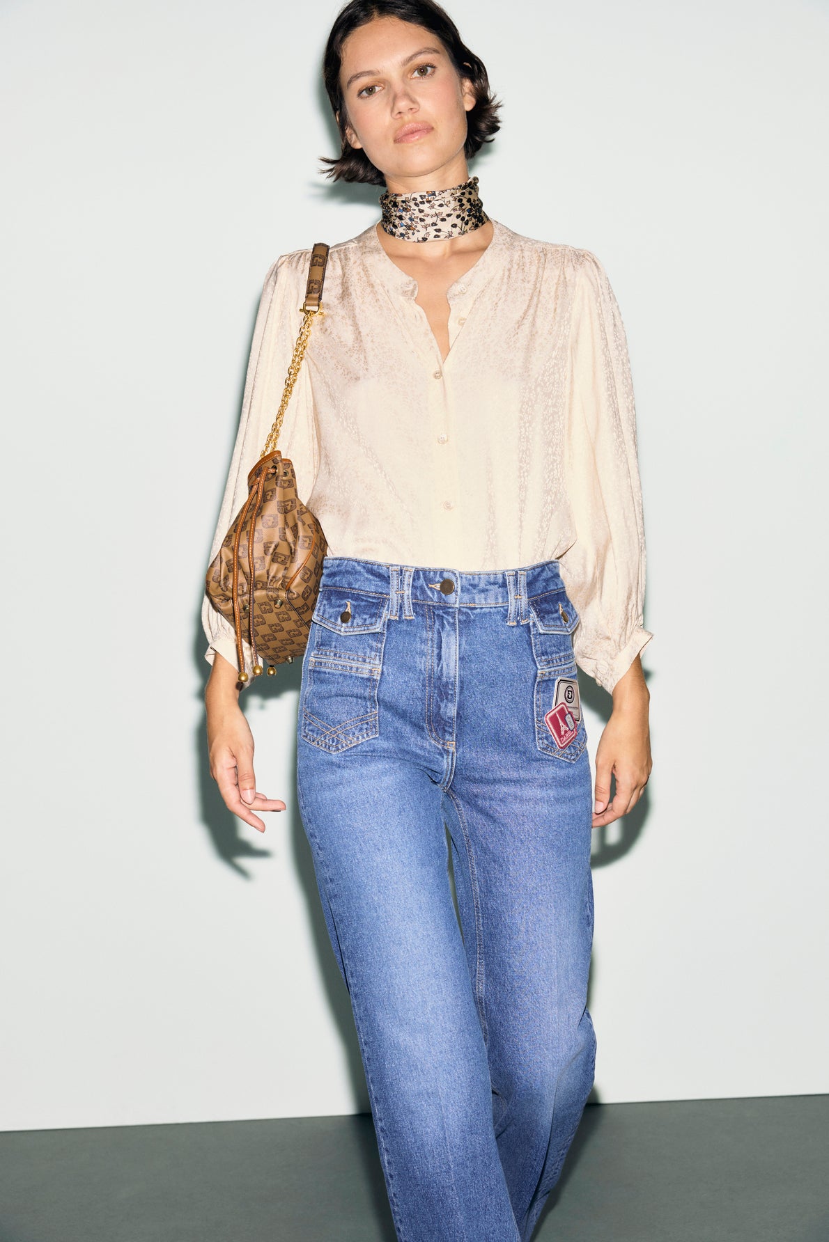 Woman wearing a cream blouse with manches bouffantes, blue high-waisted jeans with patches, a leopard print choker, and carrying a brown patterned handbag, standing against a plain background.