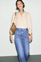 Woman wearing a cream blouse with manches bouffantes, blue high-waisted jeans with patches, a leopard print choker, and carrying a brown patterned handbag, standing against a plain background.