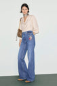 A woman wearing a light blouse with manches bouffantes, flared high-waisted jeans with patches, brown shoes, a patterned scarf around her neck, and a brown shoulder bag stands against a plain background.