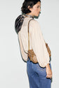 A person with short dark hair, wearing a cream chemise jacquard with manches bouffantes, a printed scarf, and blue jeans, carries a brown patterned handbag with a gold chain strap over their shoulder, standing against a plain white background.