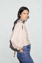 Woman with short dark hair wearing a light, long-sleeve chemise jacquard with motif floral and subtle manches bouffantes, paired with blue jeans and a printed scarf around her neck, standing sideways with one hand in her pocket against a plain light background.