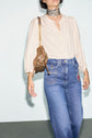 Person wearing a cream chemise jacquard with manches bouffantes, high-waisted blue jeans with patches, a patterned choker, and carrying a small brown purse with a gold chain strap, standing against a plain light background.
