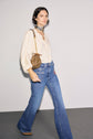 A woman wearing a cream chemise jacquard with manches bouffantes, blue high-waisted flared jeans, brown shoes, a patterned neck scarf, and a small brown shoulder bag walks indoors against a plain backdrop.