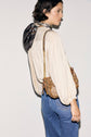 A person with short dark hair, wearing a cream chemise jacquard with manches bouffantes, a patterned scarf, and blue jeans, holds a brown chain-strap purse over their shoulder, standing against a plain light background.