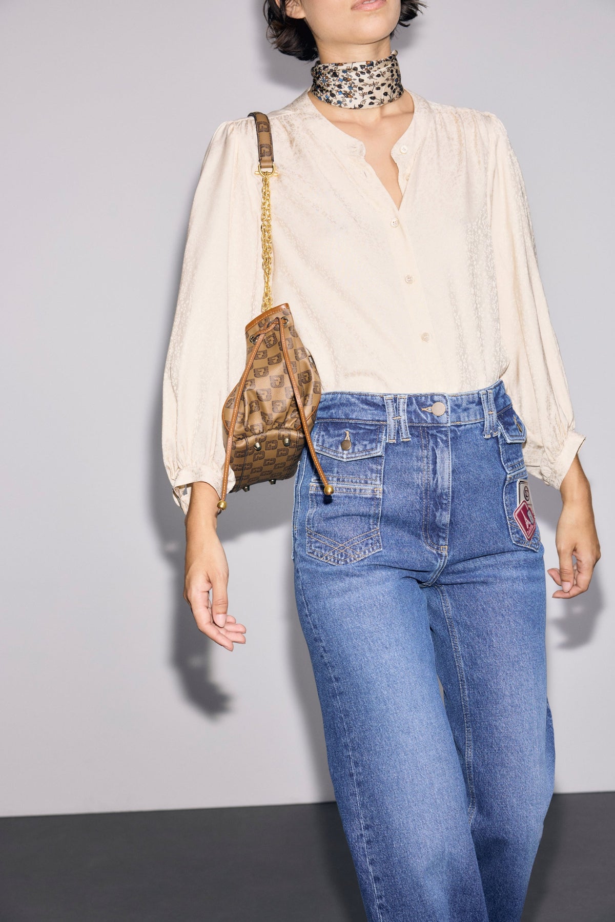 A person wearing a cream chemise jacquard with manches bouffantes, blue high-waisted jeans with patches, a printed choker, and carrying a brown patterned handbag over their shoulder stands against a plain background.
