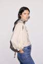 Woman with short dark hair wearing a cream-colored chemise jacquard with manches bouffantes, blue jeans, and a patterned scarf around her neck, standing sideways with one hand in her pocket against a light background.