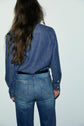 A person with long brown hair, seen from behind, wears a chemise en jean 100% coton tucked into blue jeans, standing against a plain white background, embodying an effortlessly casual chic style.