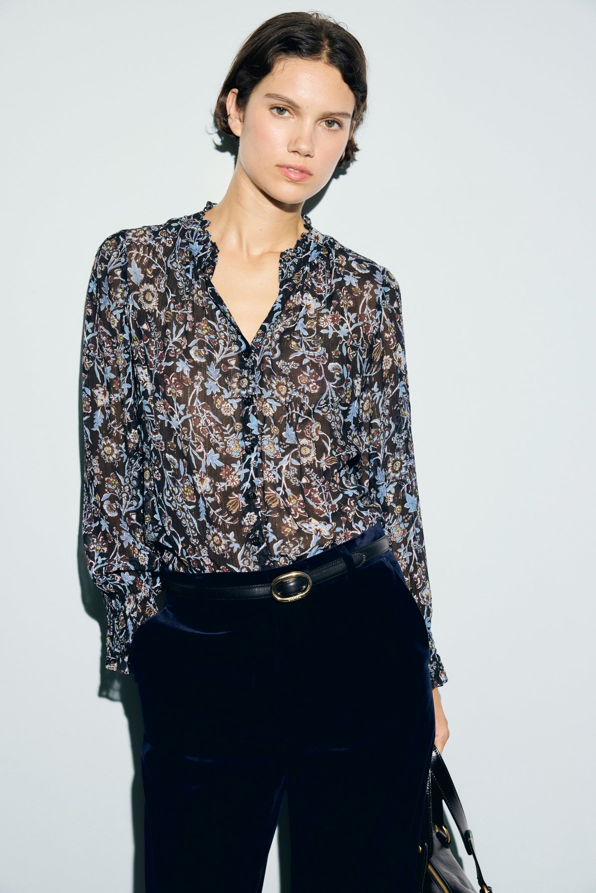 Woman with short dark hair wearing a sheer mousseline soie, floral long-sleeve blouse with col volanté and dark pants with a belt, standing against a plain light background.