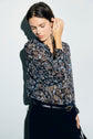 A woman wearing a dark chemise à fleurs and black pants stands against a light background, looking down and touching her hair with one hand.