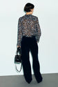 A person with short dark hair, wearing a sheer chemise à fleurs with col volanté and dark wide-leg pants, stands facing away while holding a black handbag against a plain background.