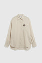 Beige, long-sleeve, button-up chemise preppy with thin vertical stripes and an embroidered dark crest on the left chest. This chemise rayée features a pointed collar and a rounded hem for a classic, polished look—a timeless chemise femme for any wardrobe.