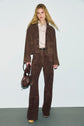 A woman with long blonde hair wears a brown suede jacket and matching flared pants, a beige chemise femme, beige platform shoes, and holds a brown leather handbag, standing against a plain light background.