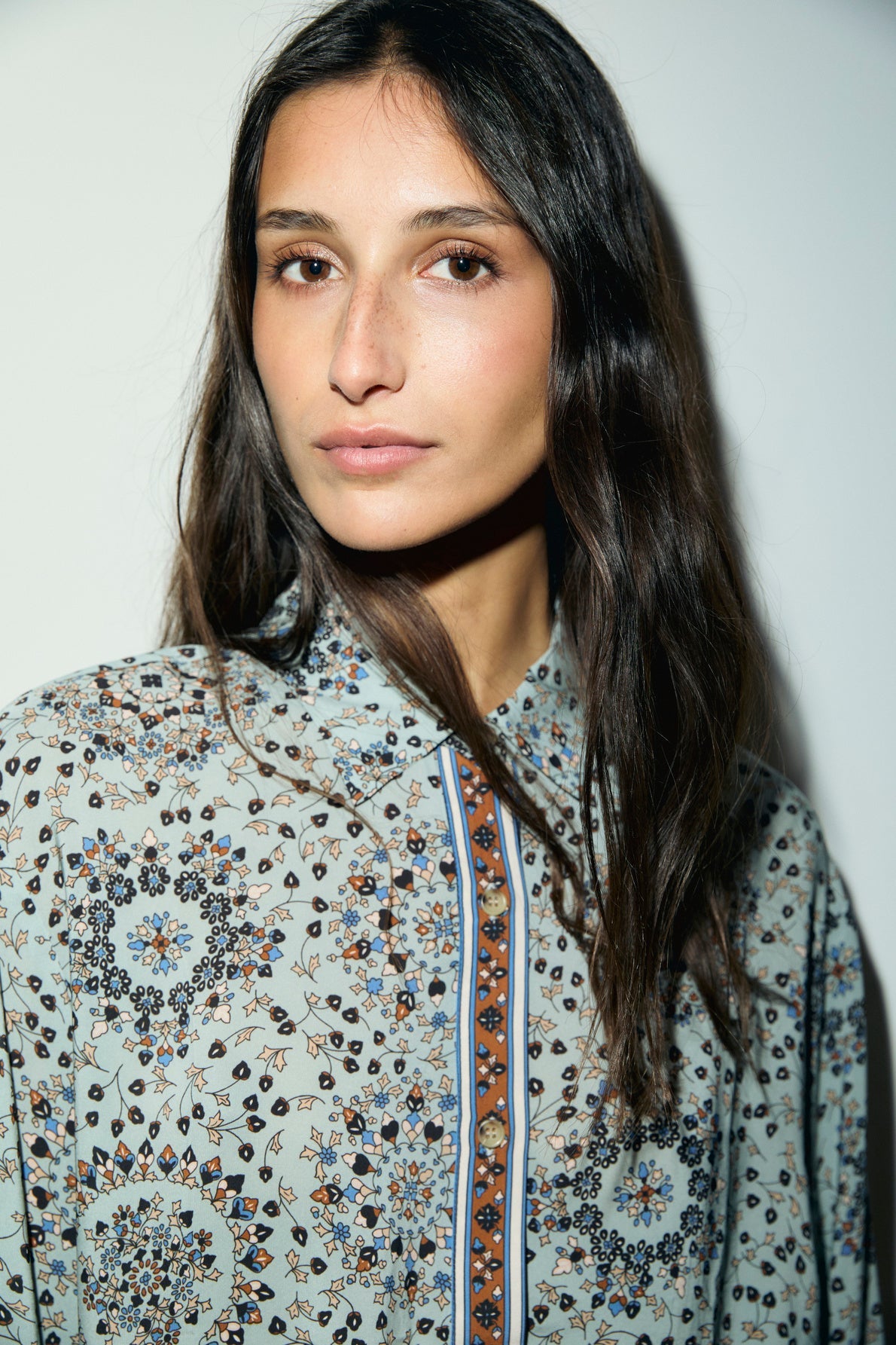 A woman with long brown hair wears a chemise fluide featuring geometric and imprimé floral designs, standing against a plain white background and looking slightly to the side.