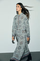 A woman with long dark hair walks, wearing a chemise fluide and wide-leg pants in muted blue and gray tones, featuring geometric and imprimé floral designs.