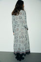 A woman with long brown hair stands facing a white wall, wearing a chemise femme à imprimé floral, long-sleeved and ankle-length, paired with black boots.