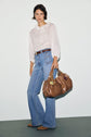 A woman stands against a plain background, wearing a chemise coton blanc with subtle volants, blue high-waisted flared jeans, a brown belt, brown shoes, and holding a large brown handbag for an effortless style bohème chic.