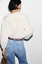A person with short dark hair wears a chemise coton blanc with volants, tucked into high-waisted blue jeans with a brown belt, holding a brown handbag, standing against a plain white background for an effortlessly style bohème chic look.