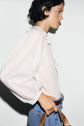 A person with short dark hair, wearing a chemise coton blanc with élégantes manches à volants and blue jeans, is holding a brown leather strap against a plain light background, capturing an effortless style bohème chic.