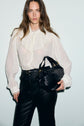 Woman with long brown hair wearing a sheer white chemise en crêpe with manches bouffantes and high-waisted black pants, holding a black leather handbag against a plain light background.