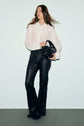 A woman stands against a light background wearing a sheer white chemise en crêpe with manches bouffantes, paired with black leather pants and holding a black handbag. Her long hair is loose, and she has a neutral expression.