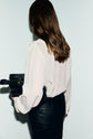 A person with long brown hair, wearing a white chemise en crêpe with manches bouffantes and black pants, is seen from behind holding a black handbag against a plain light background.