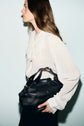 Woman in a white chemise en crêpe with delicate lace details and manches bouffantes holds a large black leather bag. She is facing left with long brown hair, against a plain light background.