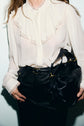 A person wears a white ruffled blouse with lace details and manches bouffantes, paired with black high-waisted pants, and holds a black leather handbag with gold hardware. Their face is partially out of frame.