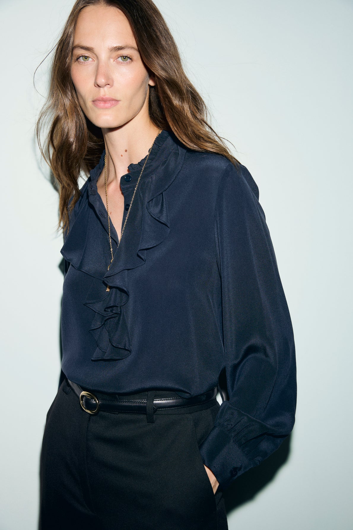 Woman with long brown hair wears a blouse en soie bleu foncé avec col volanté and black trousers, standing against a light background with one hand in her pocket, looking at the camera.
