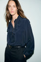 Woman with long brown hair wears a blouse en soie bleu foncé avec col volanté and black trousers, standing against a light background with one hand in her pocket, looking at the camera.